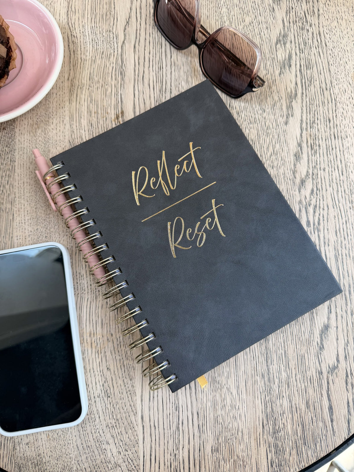 Overhead flat lay of the Reflect/Reset 2027 planner with rose gold pen on a cafe table